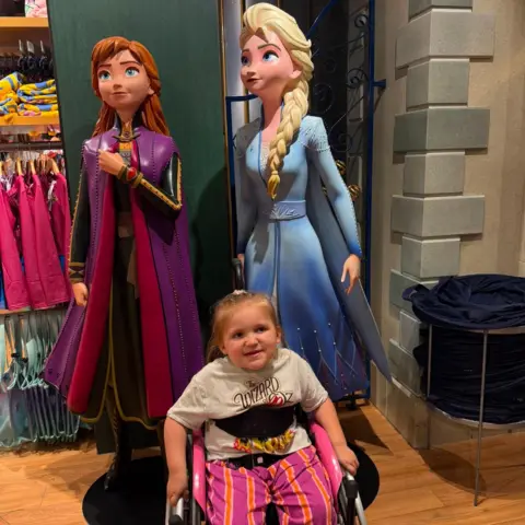 Charlie Brown A young blonde girl in a black wheelchair with black straps across her chest. She is wearing a white Wizard of Oz t shirt and pink and orange striped trousers. Behind her are life-size figures of Disney pricesses Anna and Elsa.