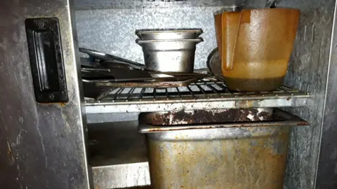 Inside a steel industrial fridge at Emma's Sandwich Shop. A plastic judge which contains a brown liquid and other metal containers are all covered in an oily, grimy layer.