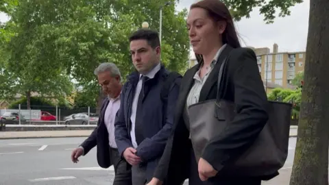 A man in a suit walks along the street accompanied by a man and a woman in business attire.