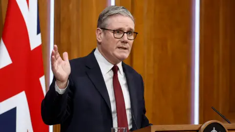 Sir Keir Starmer stands at a podium and raises his right hand. A large Union Jack is behind him.