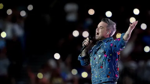Robbie Williams performs on stage in a denim jacket with colourful gems stuck all over it. He has one hand in the air with his palm up to the sky and is singing into the microphone. The crowd is blurred with circular lights visible in the background.
