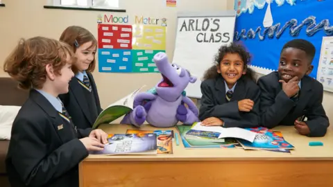 The Natasha Allergy Research Foundation Four school pupils, all wearing dark coloured blazers, light blue shirts and ties, sit round a table smiling and looking at Arlo the Armadillo, a lilac cartoonish armadillo puppet. In the background is a whiteboard with "Arlo's Class" written on it.