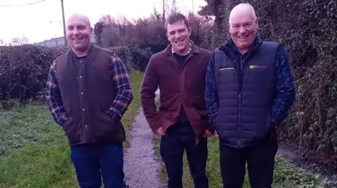 Jamie Price Three men smile on a country lane. On the right is a man in a brown gilet and checked shirt and jeans; in the middle a man in black trousers, brown jacket, black jumper and checked shirt; on the right an older man with balding grey head in navy puffa, checked navy shirt an black jeans.