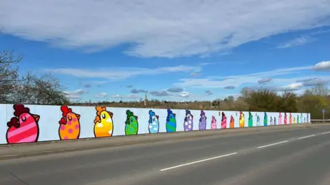 A mural on a wall at the side of a road shows paintings of chickens in various colours