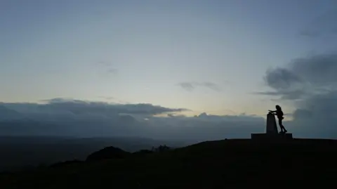 David Miller Photography A silhouette of a woman on top of a hill next to a stone. The sky behind her is blue and grey with grey clouds, and the yellow glow of the sun