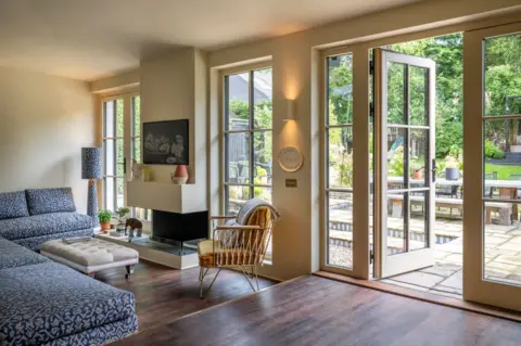 IWC Media A bright, modern living room features large glass doors and windows that open onto a green garden with a patio and outdoor seating.
Inside, a blue patterned sofa, neutral ottoman, and a sleek fireplace create a comfortable seating area with soft, natural lighting.