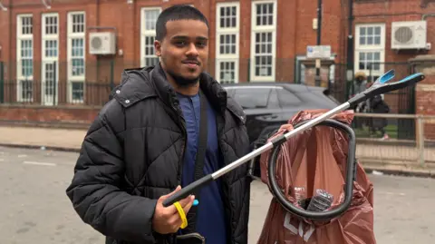 A man wearing a black puffer coat, carrying a litter picker and brown bag