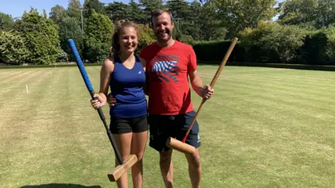 A young girl is looking at the camera and smiling in the sunshine. She has blonde hair tied back in a ponytail and is wearing a sleeveless blue vest top. Standing next to her is a man wearing sunglasses on top of his head and a red top that says Nike on it. He is also smiling and looking at the camera. Both are holding their croquet mallets. There are trees are in the background. 