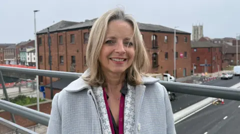 Grace McGrory/BBC A woman with long blonde hair standing on a bridge over a busy road. She is wearing a grey jacket and a patterned top.