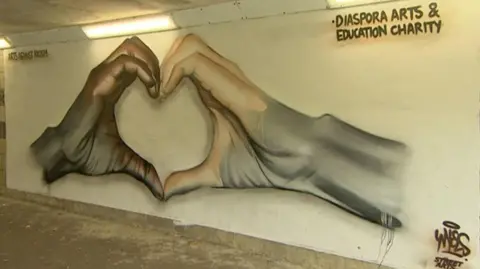 A spray-painted image of two hands coming together to form a heart shape. It is painted on the wall of an underpass - the concrete floor and roof, as well as strip lighting can be seen. The hand on the left is dark brown in colour, while the hand on the right is pink, but both hands fade into the same grey colour.