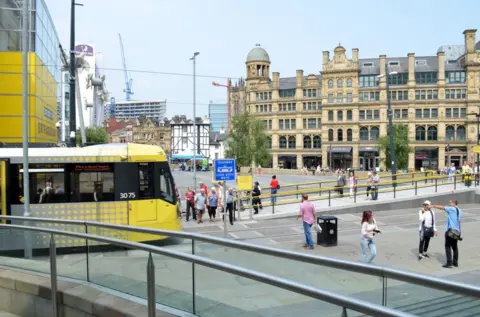 To the left, a yellow tram with dotted patterning is stopped at a platform. A warning sign near railings and a ramp with metal handrails is in the foreground. A black litter bin stands on the pavement. Pedestrians are walking across the open plaza, with historic stone buildings and construction cranes in the background.