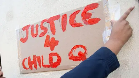 A hand places a sign on a wall reading "Justice 4 Child Q" in red paint.