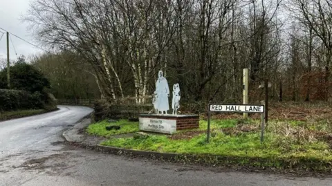 A sculpture, at the Minnie Pit memorial garden in Halmer End, situated on a grass verge at the side of a road with a sign which reads Red Hall Lane.