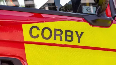 A Corby fire vehicle, in yellow and red, with a black wing mirror. 