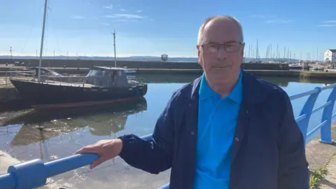 Michael Hughes stands in front of Carrickfergus marina holding the rail. He is balding and wears glasses. he has a blue polo shirt on and a navy jacket over it. It's a bright sunny day and the sky is blue. There is a little boat in the harbour behind him. 