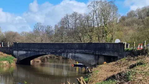 The picture shows a low, concrete road bridge crossing a calm river in a rural setting. The bridge has a single wide arch and darkened concrete, with visible staining along the sides that suggests age and weathering. Metal railings run along the top edges of the bridge, and there are a few orange-and-white traffic cones placed near one end.