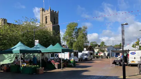 Darren Rozier/BBC A small out door market on a pedestrianised area. There are stalls under green gazebos on the right, with other vans and food outlets. There is a parish church behind the market place.