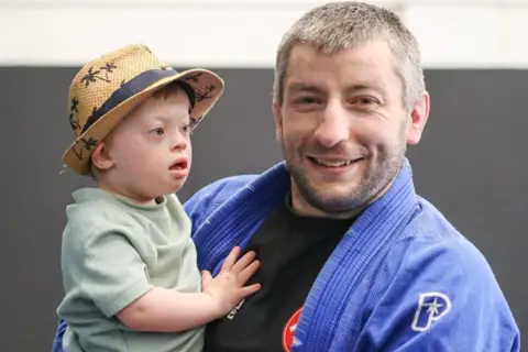 Simon Gooch A boy wearing a straw hat and green top is being held by a man with a beard wearing a blue martial arts jacket