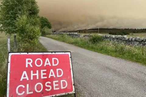 A red road warning sign set up by the side of a rural road. In white writing are the words: "Road ahead closed". There is a drystone dyke and across a field, in the distance, thick grey smoke rises from a wildfire. 