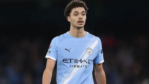 Max Alleyne during a Premier League match. He has curly brown hair and a dark brown moustache. He is wearing a Manchester City kit which is light blue and has a white diagonal stripe from one hip to one shoulder. The background is blurred and is very dark. He is lit up by bright lights.
