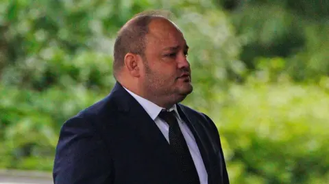 PA Media John Stringer wearing a dark suit and tie with green background