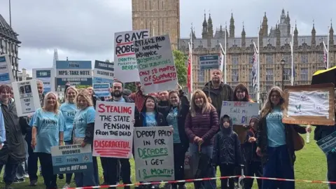 Widows Fight A group of around 20 people, a mix of men, women and children, stand on a grassy area with the UK Houses of Parliament in the background. Some are in blue t-shirts with Widows Fight on them, others are holding placards with statements about their campaign including "Daddy paid in for decades, I get 18 months"
