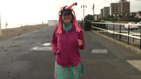 Handout A woman wearing a bright pink jacket and a green and pink dress. She is also wearing a flamingo hat while jogging along a seafront.