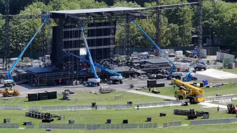 Cranes and other heavy machinery is used to set up a large stage in a wide, open park. 