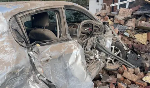 A badly damaged small car surrounded by bricks and other rubble