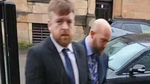 Kevin Montgomery - a man with ginger hair and a beard, arriving at court. He is dressed in a suit with a blue tie. Another man, bald and with a beard is walking beside him.
