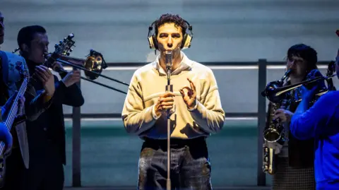Will Green Photography A production shot of Jake Halsey-Jones playing Robbie Jackson in Caroline. He is talking into a microphone which is on a stand and is wearing a pair of headphones. He is wearing a beige jumper which has a white shirt underneath and a pair of jeans. Around him are the rest of the cast playing instruments including the trumpet, trombone and saxophone.