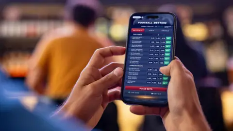Getty Images A man holding a smartphone with betting odds displayed on the screen.
