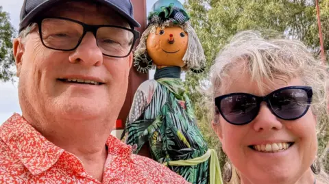 Stephen Howe Stephen How, pictured left, wearing an orange floral short. He is standing next to his wife Elizabeth Howe, who is wearing dark sunglasses. There is a scarecrow behind them.