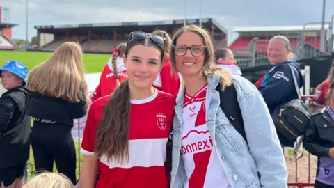 Two Hull KR fans smiling for a photo at a rugby ground, wearing red and white team shirts, with other supporters and stadium seating in the background.