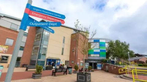 An exterior view of the University Hospital of North Durham, a brick building with blue and green panels and glass windows. In the courtyard of the building there is a signpost with directions to different hospital departments.
