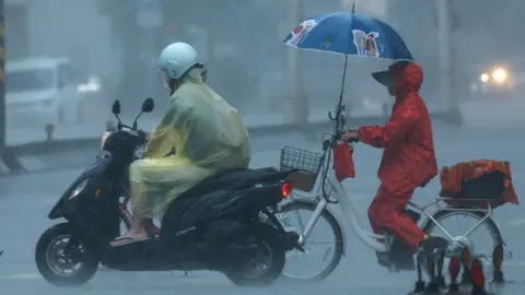 Cyclists sit in heavy rain due to Typhoon Dolphin in Taiwan. One wears a yellow cagoule and the other holds a blue umbrella.