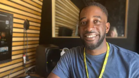 A man smiles as he sits in a wood panelled music studio. He is wearing a blue top and a blue T-shirt. He has short black hair.
