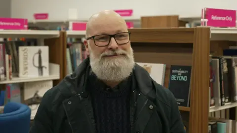 A man with a white beard and wearing glasses is in a library