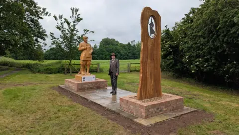 Wooden sculpture of JRR Tolkien depicted in his army uniform and holding a notebook, along with a second sculpture depicting the writer's wife Edith danncing. There is a man standing between the two structures dressed in a suit and hat.