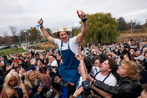 AP Ben Shabad, the winner of the Jeremy Allen White lookalike contest, wearing a chef's apron and white t-shirt, is hoisted on the shoulders of crowd members in a Chicago park