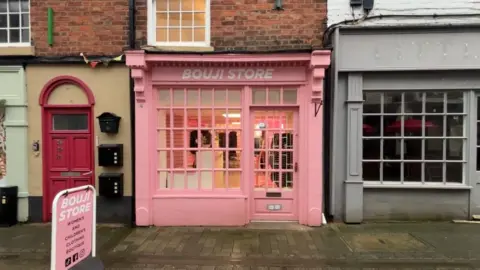 BBC/ Alex James A light pink store front with the door to the right of the shop window. The shop name is written in white text above the door and window and reads Bouji Store. There is a grey empty shop front on the right of the photo and a red front door on the left of it. 