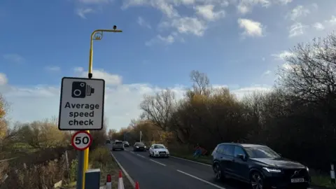 Andrew Turner/BBC A speed limit sign bearing the numeral 50, in a red circle. Above that is another sign, showing a pictogram of a camera, with the text "average speed check" within, and behind that, a yellow standard which arcs to the right, upon which is a camera. Traffic can be seen in both directions on a single-carriageway road. Hedges can be seen either side of the road.