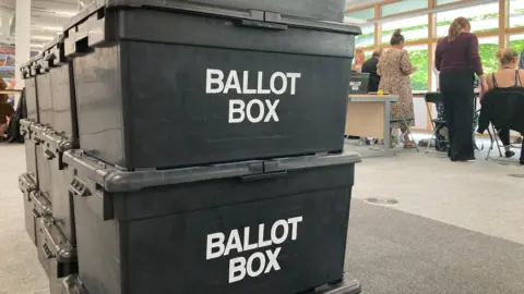There are a pile of black ballot boxes stacked inside a counting centre. A number of people can be seen in the background sitting and standing at desks.