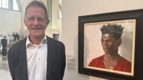 Wearing a grey a suit over an open neck white shirt, a smiling Sir Nicholas Serota is also wearing a pair of round, almost rimless glasses. He's standing alongside a portrait of the artist Nahem Shoa.