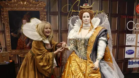 A woman, with blonde hair, wearing an Elizabethan-era costume stands next to the life-size cake of Queen Elizabeth I