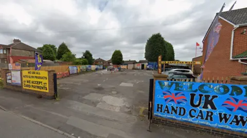 Google A colourful blue car wash sign can be seen on the right of the entrance to the land with a yellow card payments sign visible on the left side. There is a large space once through the entrance to the car wash. There is a concrete ground and the car wash is on the right side towards the back. There is low-level fencing around the site.