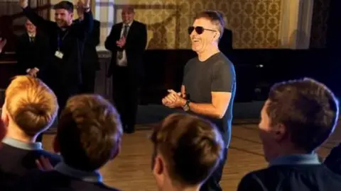 Simon Cowell stands to the right of the boys who are in a semi circle gathered around him laughing and cheering, some seen from the back. They were navy school jumpers and light blue polo shirts