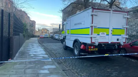 BBC A bomb disposal van parked on High School Yards in Edinburgh. The van is white, with a yellow and blue stripe round it. It has the words Bomb Disposal in black lettering on the side. A thin line of white and blue police tape is in the foreground.