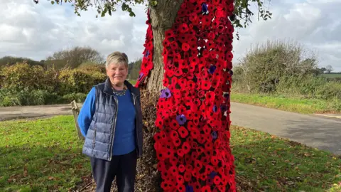 Clare Worden/BBC Woman stands in front of a tree which is covered in a cascade of knitted poppies in various shades of red and purple. The woman is smiling and wears a navy blue gilet over a royal blue jumper and a necklace.