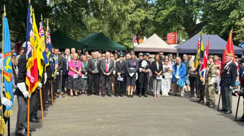BBC People are holding flags of various colours on the far left and far right of the photo. In between them, dozens of people are in the background facing the camera. 